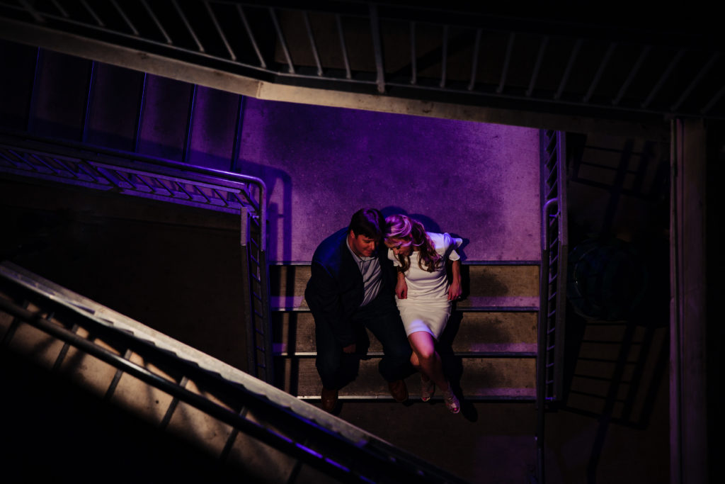 Colorful night time portrait of engaged couple cuddling on stairs in The Pearl Historic District in San Antonio, TX. Photography by Shannon Cain Photography