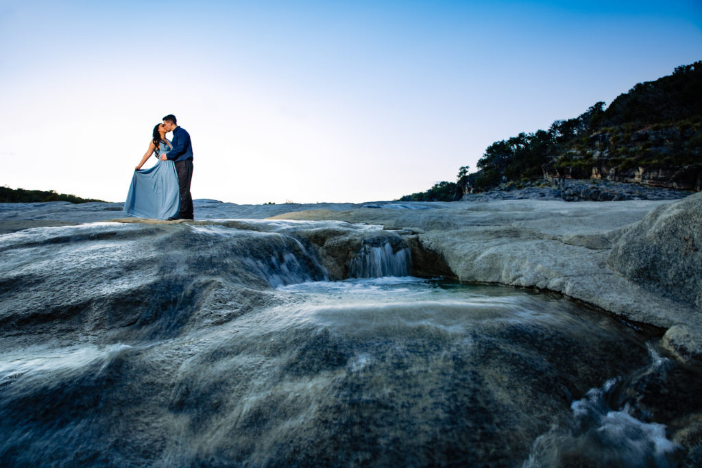 Couple pose for engagement portrait at Pedernales Falls State Park by water flowing over the falls in formal outfits.  Photography by Shannon Cain Photography