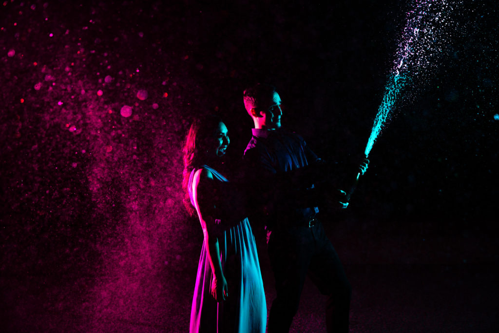 Couple celebrating their engagement with a colorful night time champagne bottle pop. Photography by Shannon Cain Photography