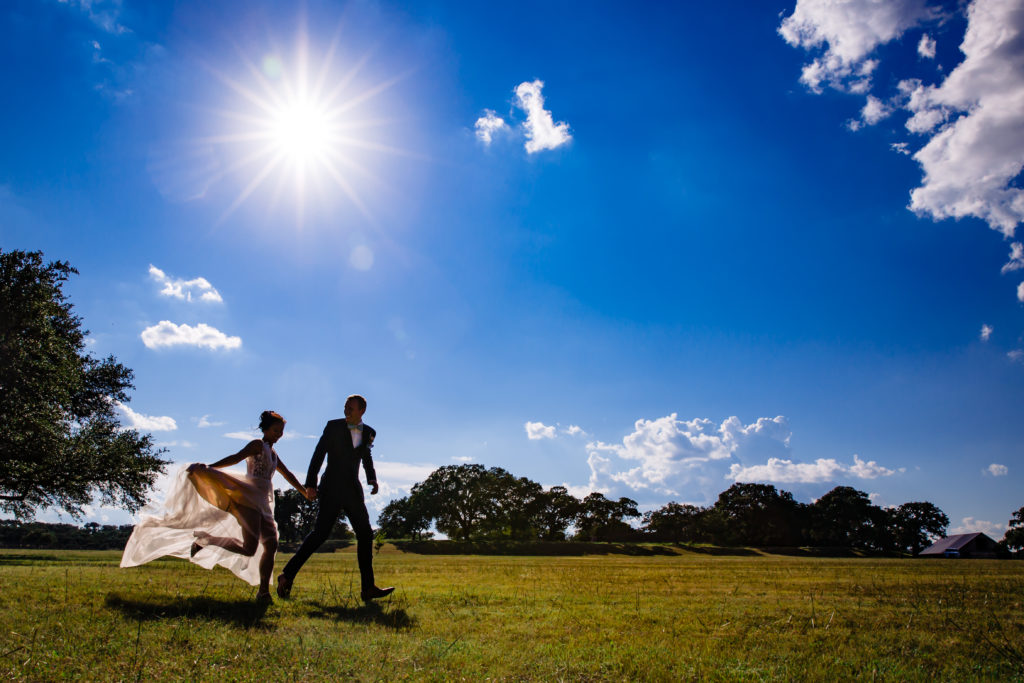 Bride and groom hold hands and run across a field together with the sun shining on them at Bergheim Ranch in Boerne.  Photo by Shannon Cain Photography