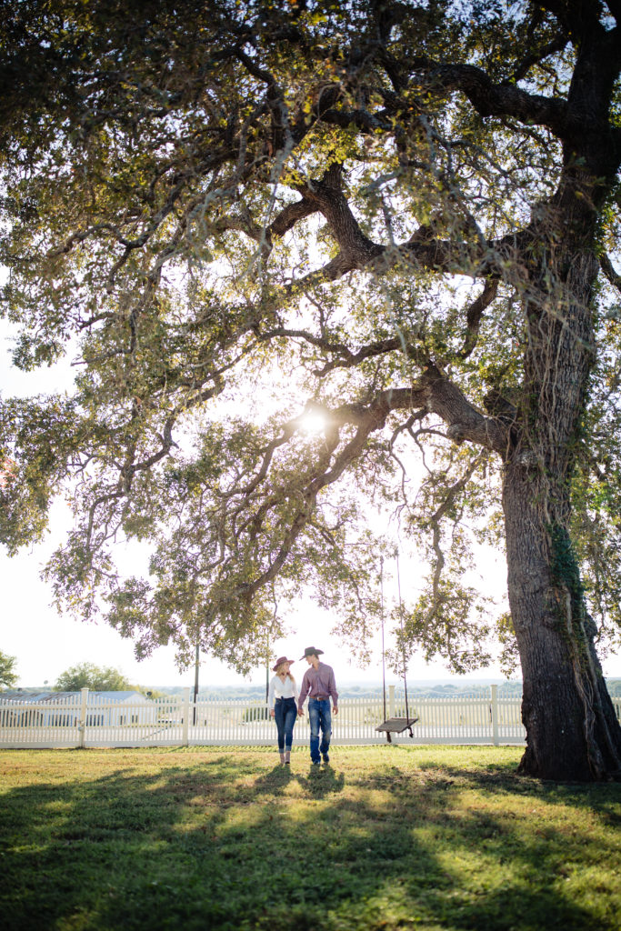 Cute couple stroll in jeans and hats for engagement photos under a big oak tree with a swing at Whitehall Polley Mansion in La Vernia, TX. Photography by Shannon Cain Photography
