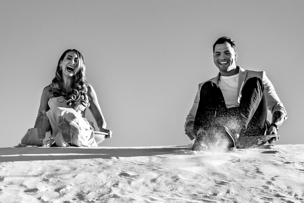 Bride and Groom sledding at The White Sands National Park at sunset for Adventure Engagement Session.  Photography by Shannon Cain Photography.  Hair and Makeup by Glam on Demand.